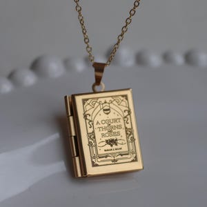 Può includere: Collana con pendente a forma di libro color oro. Il pendente è inciso con il testo "A Court of Thorns and Roses" e il nome dell'autore. La collana ha una catena delicata.