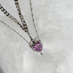 May include: A silver necklace featuring a heart-shaped pendant with a pink gemstone center, surrounded by clear crystals. The pendant is pierced by a dagger. The necklace has two chain styles: a chunky curb chain and a delicate cable chain.