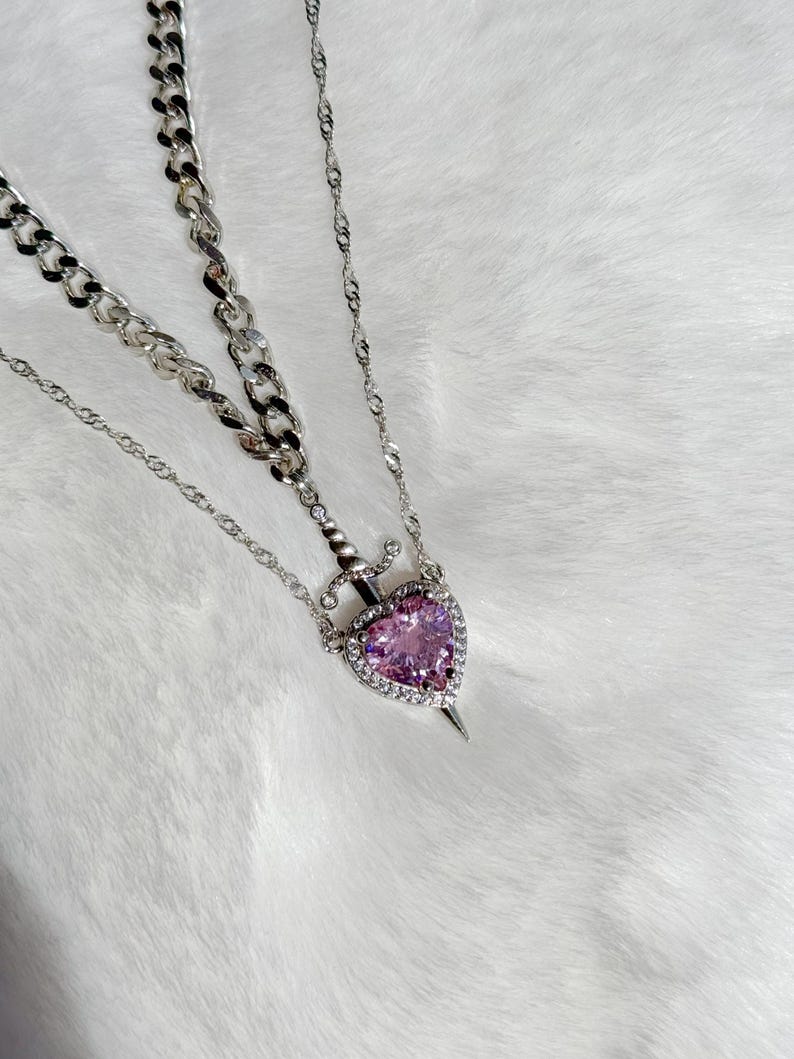 May include: A silver necklace featuring a heart-shaped pendant with a pink gemstone center, surrounded by clear crystals. The pendant is pierced by a dagger. The necklace has two chain styles: a chunky curb chain and a delicate cable chain.