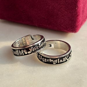 Könnte beinhalten: Zwei silberne Ringe mit schwarzer Schrift. Auf einem Ring steht "will lift your soul" und auf dem anderen "this ring lasts". Die Ringe liegen auf einer weißen Oberfläche mit einer weinroten Schachtel im Hintergrund.