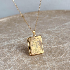 Puede incluir: Un collar de cadena dorada con un colgante de medallón que presenta un diseño de carta de tarot. La carta representa a una pareja con el texto "THE LOVERS" debajo.