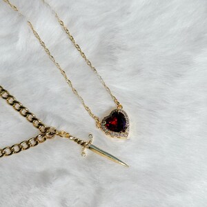May include: Gold-toned necklaces with pendants. One necklace features a heart-shaped pendant with a deep red stone and a halo of small clear stones. The other necklace has a dagger pendant with a small clear stone accent.