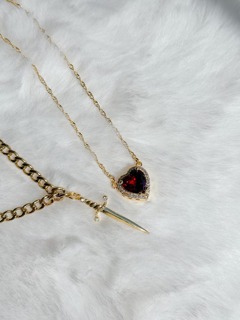 May include: Gold-toned necklaces with pendants. One necklace features a heart-shaped pendant with a deep red stone and a halo of small clear stones. The other necklace has a dagger pendant with a small clear stone accent.