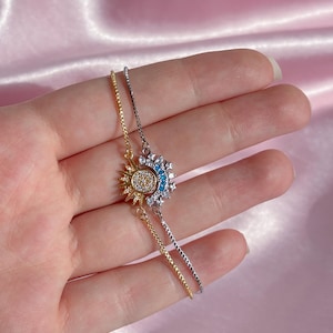 Può includere: Un braccialetto delicato con un ciondolo a forma di sole e luna. Il ciondolo a forma di sole è dorato con cristalli trasparenti, mentre la luna è argentata con cristalli blu. Il braccialetto ha una catena sottile ed è probabilmente in metallo.