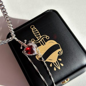 May include: A silver necklace featuring a pendant with a red heart-shaped gemstone, surrounded by smaller clear stones. The pendant is designed as a sword piercing the heart. The necklace rests on a black box with a gold heart and dagger design.