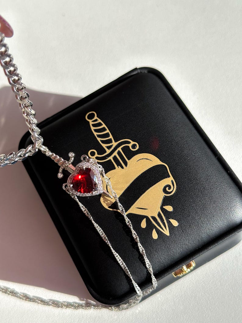 May include: A silver necklace featuring a pendant with a red heart-shaped gemstone, surrounded by smaller clear stones. The pendant is designed as a sword piercing the heart. The necklace rests on a black box with a gold heart and dagger design.