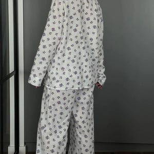 May include: White pajama set with a floral pattern in shades of purple. The set includes a long-sleeved button-down shirt and wide-leg pants. The fabric appears to be lightweight and comfortable.