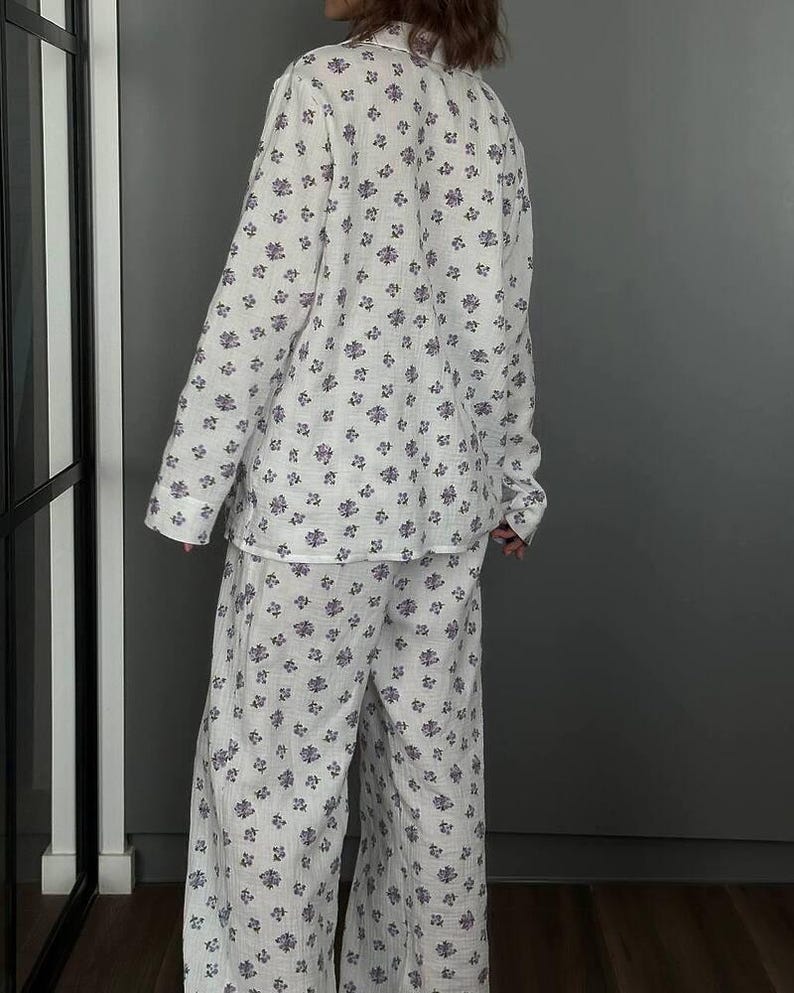 May include: White pajama set with a floral pattern in shades of purple. The set includes a long-sleeved button-down shirt and wide-leg pants. The fabric appears to be lightweight and comfortable.