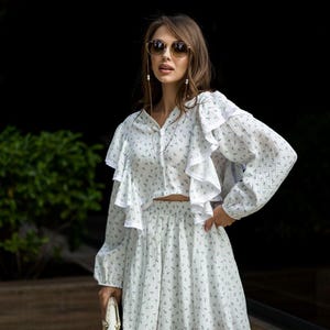 May include: A two-piece white floral print outfit with a ruffled top and a matching skirt. The top has long sleeves and a button-down front. The skirt is mini length. The outfit is paired with sunglasses and a white handbag.
