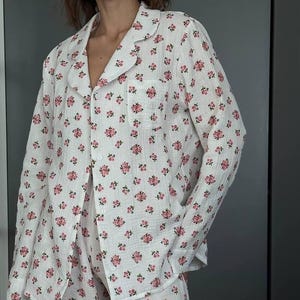 May include: White pajama set with a pink floral pattern. The set includes a button-down shirt with a chest pocket and matching pants. The fabric appears lightweight and comfortable, ideal for sleepwear.