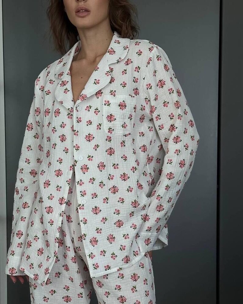 May include: White pajama set with a pink floral pattern. The set includes a button-down shirt with a chest pocket and matching pants. The fabric appears lightweight and comfortable, ideal for sleepwear.