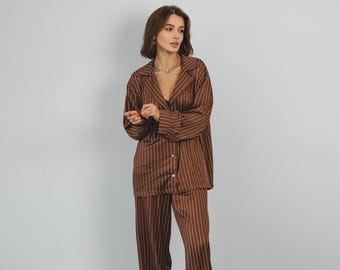 Puede incluir: Conjunto de pijama a rayas marrones. El conjunto incluye una camisa con botones y pantalones de pernera ancha. El pijama está hecho de un material suave y sedoso. La modelo lleva un collar dorado y zapatillas grises peludas.