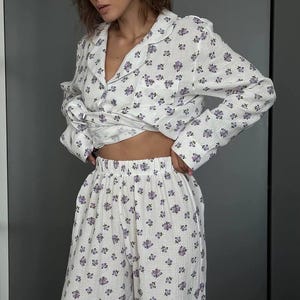 May include: White pajama set with a delicate purple floral pattern. The set includes a button-down shirt with a notched collar and long sleeves, and matching wide-leg pants. The fabric appears soft and lightweight, perfect for sleepwear.