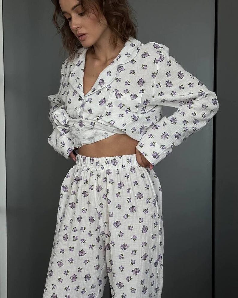 May include: White pajama set with a delicate purple floral pattern. The set includes a button-down shirt with a notched collar and long sleeves, and matching wide-leg pants. The fabric appears soft and lightweight, perfect for sleepwear.