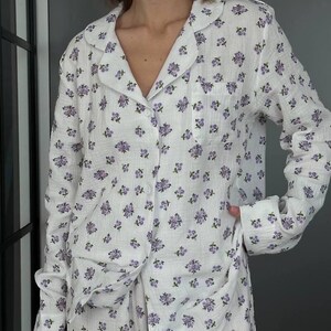 May include: White pajama set with a delicate purple floral pattern. The long-sleeved button-down shirt has a notched collar and a chest pocket. The matching pants are visible at the bottom. The fabric appears soft and lightweight, ideal for sleepwear.