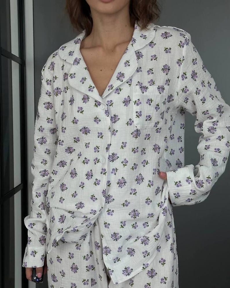 May include: White pajama set with a delicate purple floral pattern. The long-sleeved button-down shirt has a notched collar and a chest pocket. The matching pants are visible at the bottom. The fabric appears soft and lightweight, ideal for sleepwear.