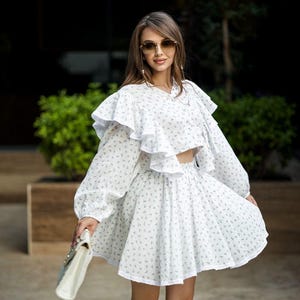 May include: White two-piece outfit with a floral pattern. The set includes a long-sleeved crop top with ruffles and a matching mini skirt. A white handbag is held in the hand. The outfit is worn with sunglasses.