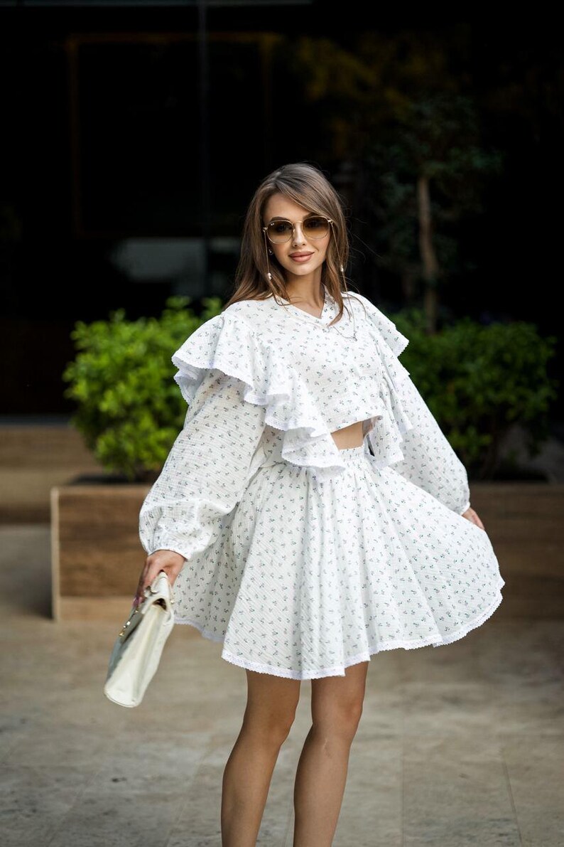 May include: White two-piece outfit with a floral pattern. The set includes a long-sleeved crop top with ruffles and a matching mini skirt. A white handbag is held in the hand. The outfit is worn with sunglasses.