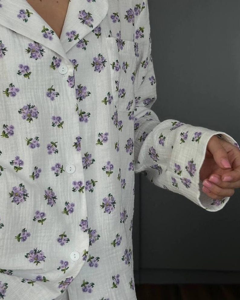 May include: White pajama set with a delicate floral pattern in shades of purple and green. The long-sleeved top features a button-down front and a chest pocket. The fabric appears soft and lightweight, ideal for sleepwear.