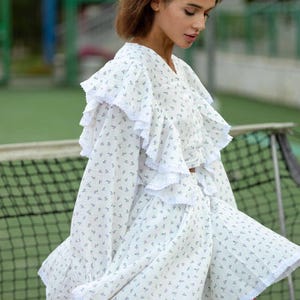 May include: White floral print dress with ruffled sleeves and hem. The dress features a V-neckline and a delicate pattern of small blue flowers. The fabric appears lightweight and airy, suitable for warm weather.