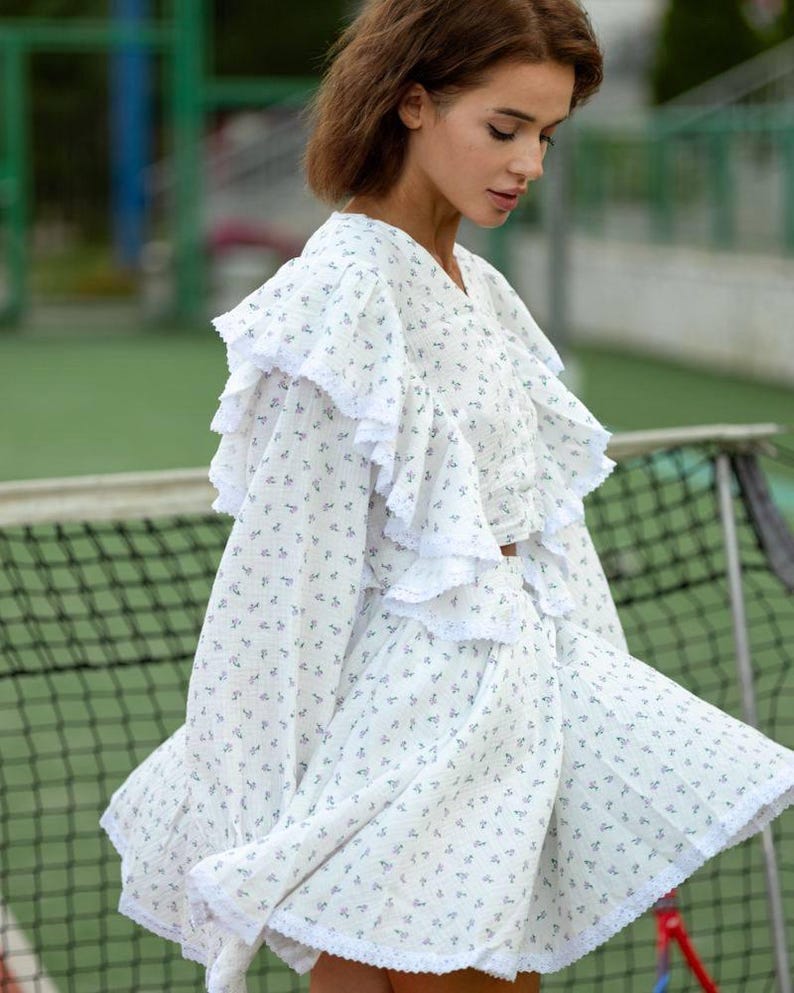 May include: White floral print dress with ruffled sleeves and hem. The dress features a V-neckline and a delicate pattern of small blue flowers. The fabric appears lightweight and airy, suitable for warm weather.
