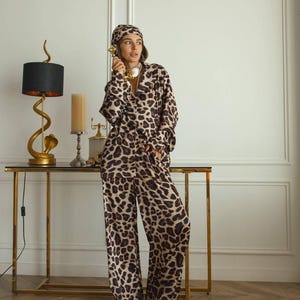 May include: A leopard print pajama set, comprising a button-down shirt, wide-leg trousers, and a headscarf. The set is in brown and tan tones. The outfit is worn with a gold necklace and a vintage gold telephone.
