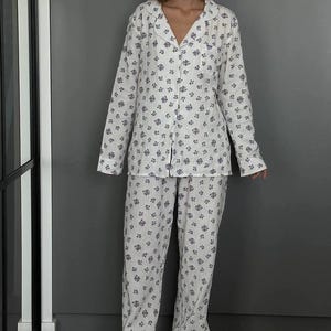 May include: White pajama set with a blue floral pattern. The set includes a button-down shirt with a notched collar and long sleeves, and matching pants. The pajamas are made of a lightweight fabric, perfect for lounging. The model is wearing gray slippers.
