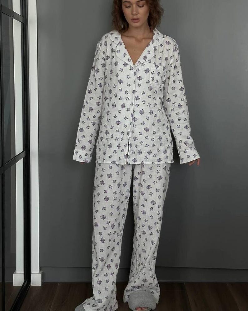 May include: White pajama set with a blue floral pattern. The set includes a button-down shirt with a notched collar and long sleeves, and matching pants. The pajamas are made of a lightweight fabric, perfect for lounging. The model is wearing gray slippers.