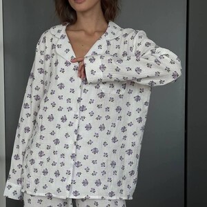 May include: White pajama set with a delicate floral pattern in shades of purple. The set includes a button-down shirt with a collar and a pair of matching pants. The fabric appears soft and lightweight, perfect for comfortable sleepwear.