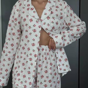 May include: White pajama set with a pink floral pattern. The set includes a button-down shirt with a chest pocket and matching shorts. The fabric appears to be a lightweight, textured material.