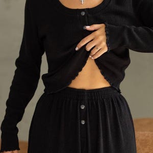 Pode incluir: Conjunto de roupa de descanso preto de duas peças. O conjunto inclui um top de manga comprida com botões e calças a condizer. O top tem decote redondo e bordas onduladas. As calças têm fecho de botão. O tecido parece ser malha texturizada.