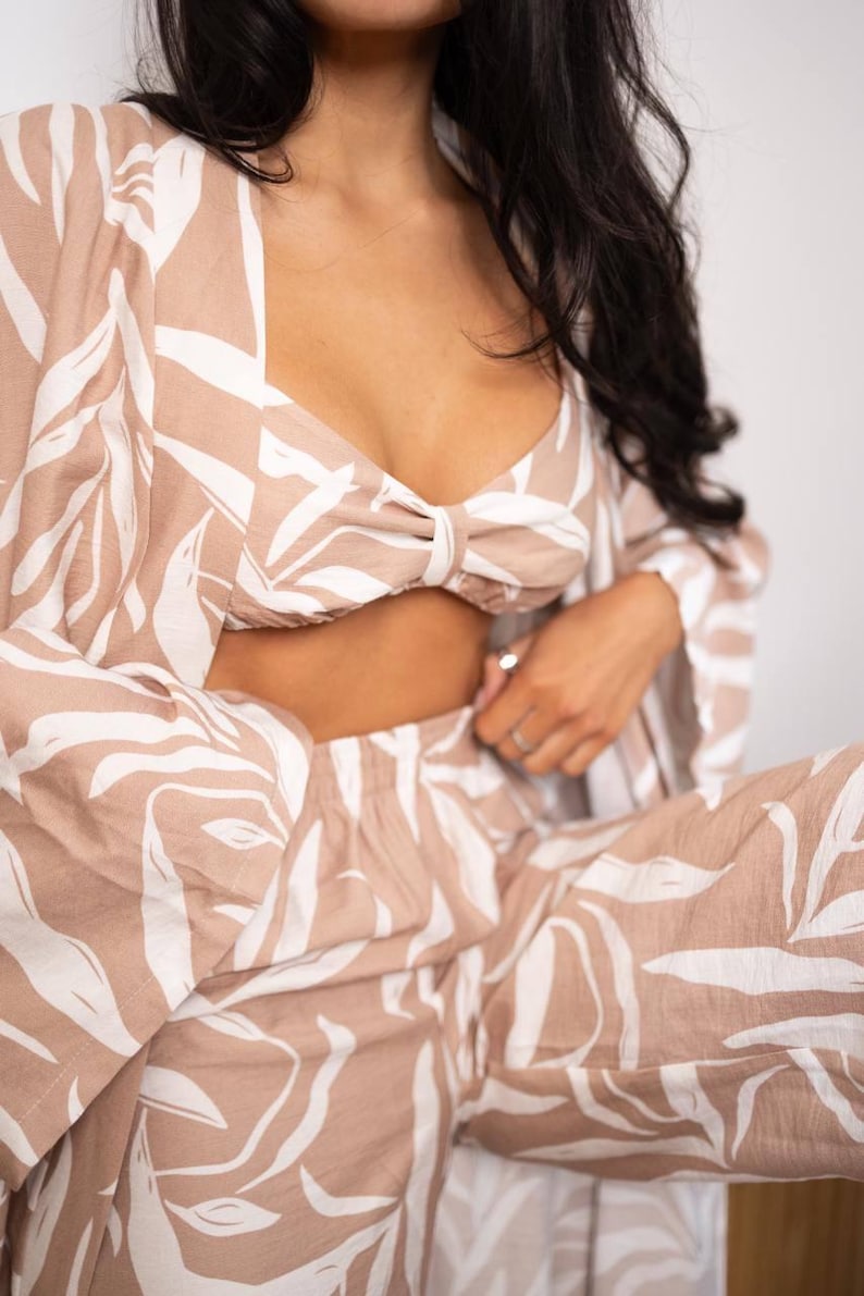 Puede incluir: Conjunto de tres piezas con estampado de hojas beige y blanco. El conjunto incluye un top corto con detalle de lazo, una chaqueta tipo kimono de manga larga y pantalones a juego. La tela tiene un aspecto suave y fluido.