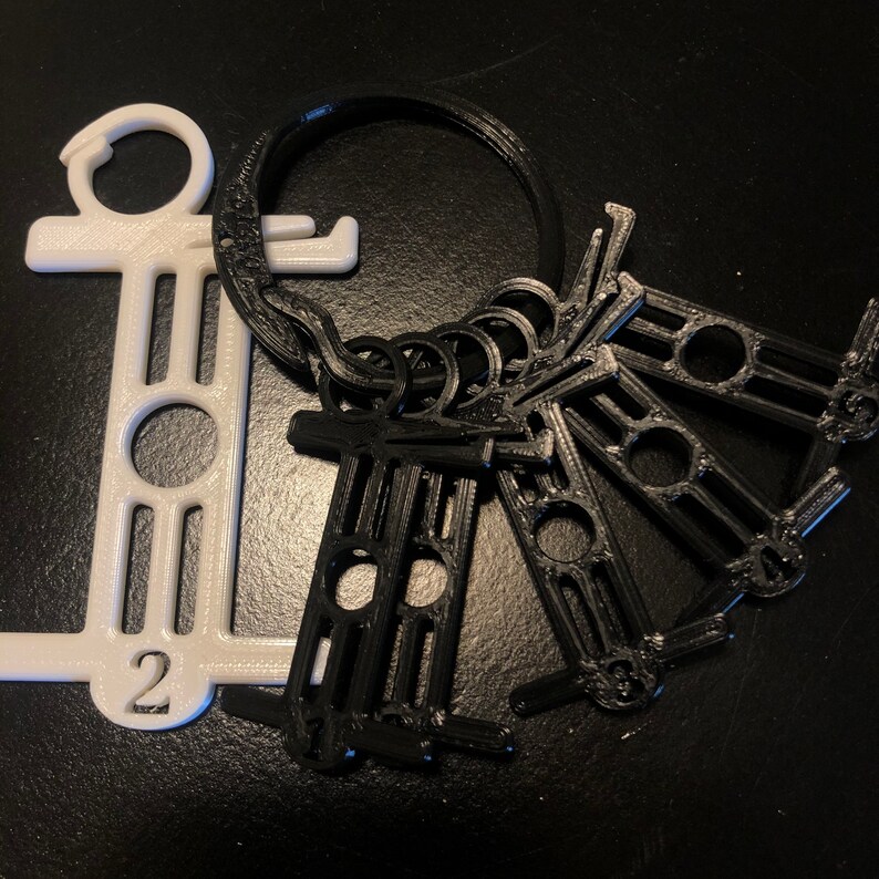 May include: A collection of 3D-printed keychains. One white keychain with the number 2, and several black keychains attached to a ring. The keychains have a unique design with circular and rectangular cutouts.