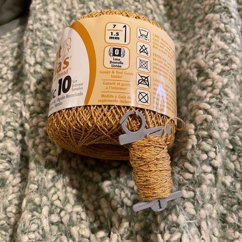 May include: A spool of gold metallic crochet thread with a size 10 label and a 1.5 mm thickness. A gray plastic thread guide is attached. The thread is on a textured, light green surface.