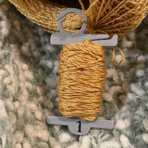 May include: A spool of gold yarn with metallic flecks, held by a gray plastic holder. The holder has a number 1 printed on it. The yarn is set against a textured, light green and white background.