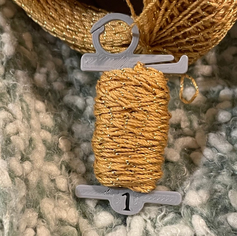May include: A spool of gold yarn with metallic flecks, held by a gray plastic holder. The holder has a number 1 printed on it. The yarn is set against a textured, light green and white background.