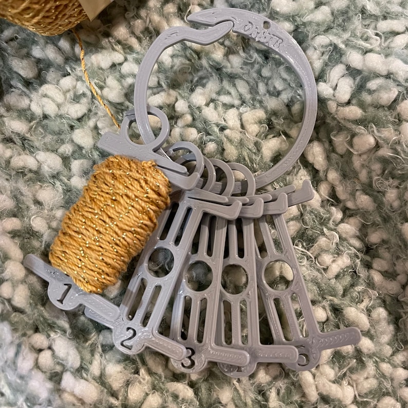 May include: A gray 3D-printed yarn gauge with a ring and several numbered measuring tools. A small spool of gold-colored yarn is attached. The word "Dohm" is printed on the ring. The numbers 1, 2, and 3 are visible on the measuring tools.