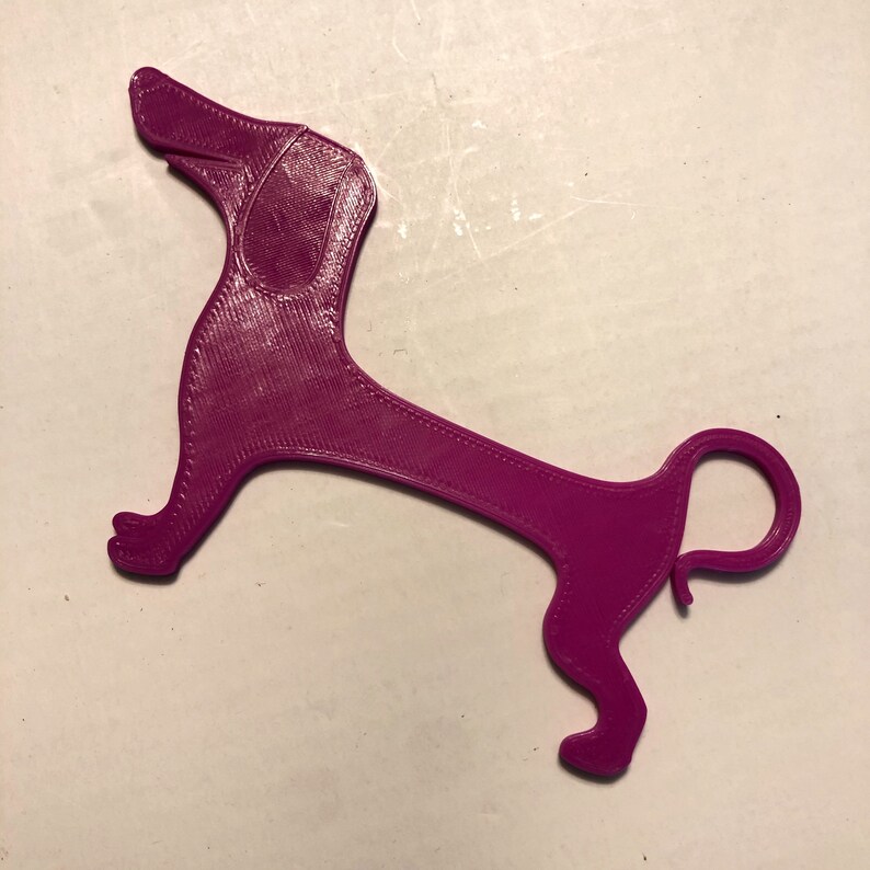 May include: A 3D printed purple dachshund dog bookmark. The bookmark is shaped like a dachshund dog with a long body and short legs. The bookmark is made of a smooth, glossy plastic.