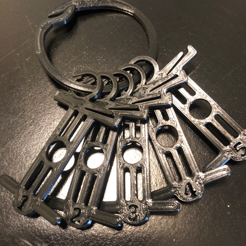 May include: Black 3D-printed keychains with a circular ring and several numbered key tags. Each tag has a unique number from 1 to 5. The keychains are made of a dark, matte material.