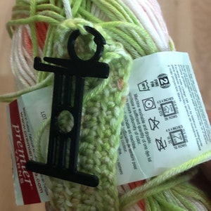 May include: A close-up of a skein of yarn in shades of green, white, and orange, held together with a black plastic yarn holder. The yarn label provides washing instructions and gauge information.