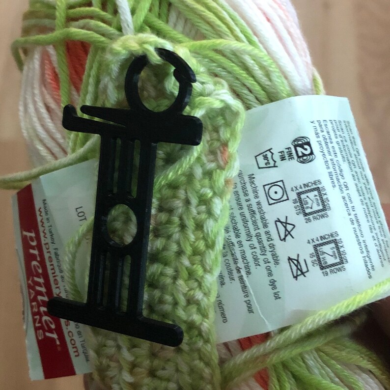 May include: A close-up of a skein of yarn in shades of green, white, and orange, held together with a black plastic yarn holder. The yarn label provides washing instructions and gauge information.
