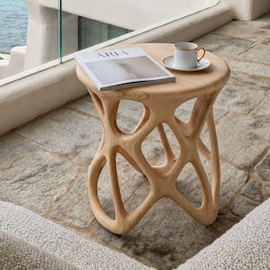 May include: A light-colored wooden side table with a sculptural, organic base featuring interconnected, curved supports. A white ceramic coffee cup on a matching saucer and a magazine with the word ARIA on the cover rest on the round tabletop.