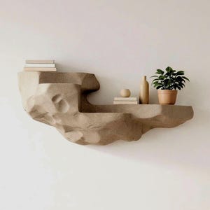 Puede incluir: Un estante flotante montado en la pared con una apariencia texturizada similar a la roca en color beige claro. El estante cuenta con dos niveles que sostienen una pila de libros, un objeto esférico pequeño, un jarrón cilíndrico y una planta verde en maceta.