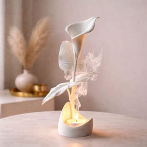 Puede incluir: Un portavelas de cerámica blanca con forma de flor de cala. Tiene una base con una vela encendida, y la flor y las hojas están encima. El humo se eleva de la vela, creando un ambiente sereno y elegante.