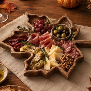 Op de afbeelding: Een houten serveerplank in de vorm van een esdoornblad, gevuld met een assortiment charcuterie waaronder prosciutto, salami, groene olijven, artisjokharten, zongedroogde tomaten, kaasblokjes en walnoten, gerangschikt op een linnen doek.