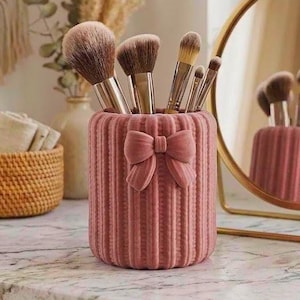May include: A cylindrical makeup brush holder with a dusty rose velvet texture, featuring vertical ribbed detailing and a matching velvet bow on the front. The holder contains five various makeup brushes with metallic handles and soft bristles.