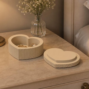 May include: A heart-shaped beige trinket box with a ribbed exterior texture. The box consists of a base and a separate lid, both featuring a consistent vertical fluted pattern along the sides. The interior of the base is smooth and beige.