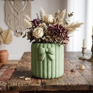 Può includere: Un vaso in ceramica verde salvia con un dettaglio a fiocco, riempito di fiori secchi nei toni crema, bordeaux e beige. Il vaso ha una trama intrecciata e poggia su un tavolo di legno rustico, creando un'estetica rustica.