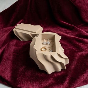 Può includere: Un portagioie beige aperto, con coperchio, su uno sfondo di velluto rosso scuro. All'interno, la scatola contiene un anello d'oro, una delicata collana e un paio di orecchini ornati. La scatola ha una forma organica unica.
