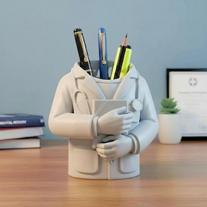 May include: A white desk organizer shaped like a doctor, holding a book. The organizer holds pens and a pencil. The pens are black and blue. The pencil is yellow and black. The organizer is on a wooden desk.